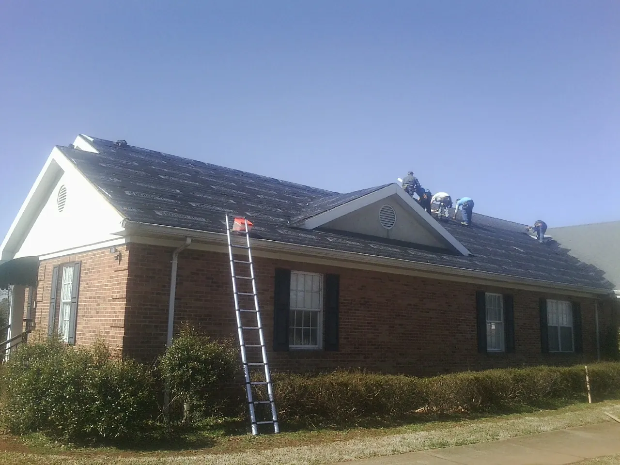 Crew tearing shingles off a brick ranch roof with a ladder set — Emergency Roof Repair work by our crew in La Rose