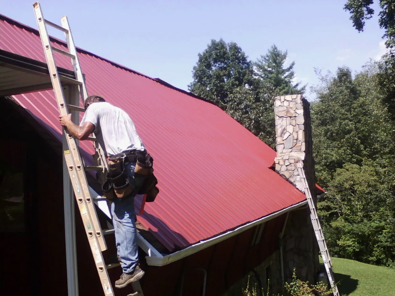 Roofer on a ladder beside a red metal roof and stone chimney — Soffit & Fascia Repair by Roofing Brothers in La Rose